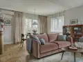 Przytulny salon z różową kanapą na środku, stolikiem kawowym, regałem i jadalnią w tle.