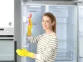 Kobieta w żółtych rękawiczkach czyści wnętrze lodówki różową gąbką. Pokazuje, jak myć lodówkę, by była lśniąca.