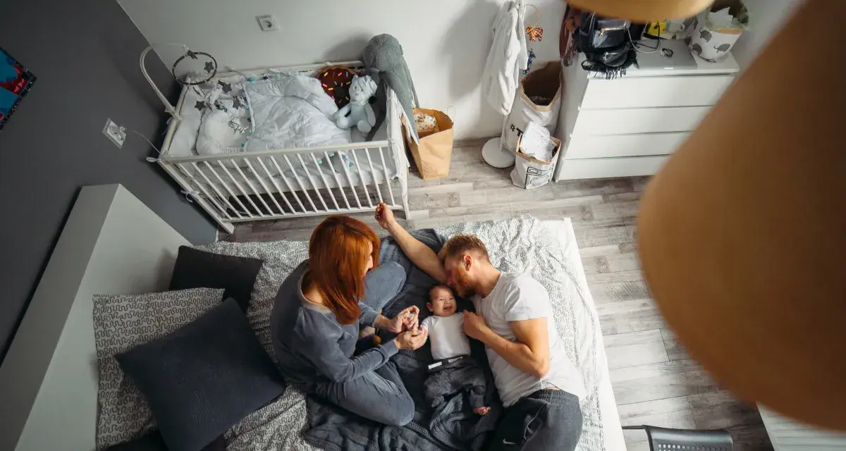 Rodzice bawią się z dzieckiem na łóżku w pokoju, który jest jednocześnie sypialnią i pokojem dziecka. Obok łóżeczko dziecięce.