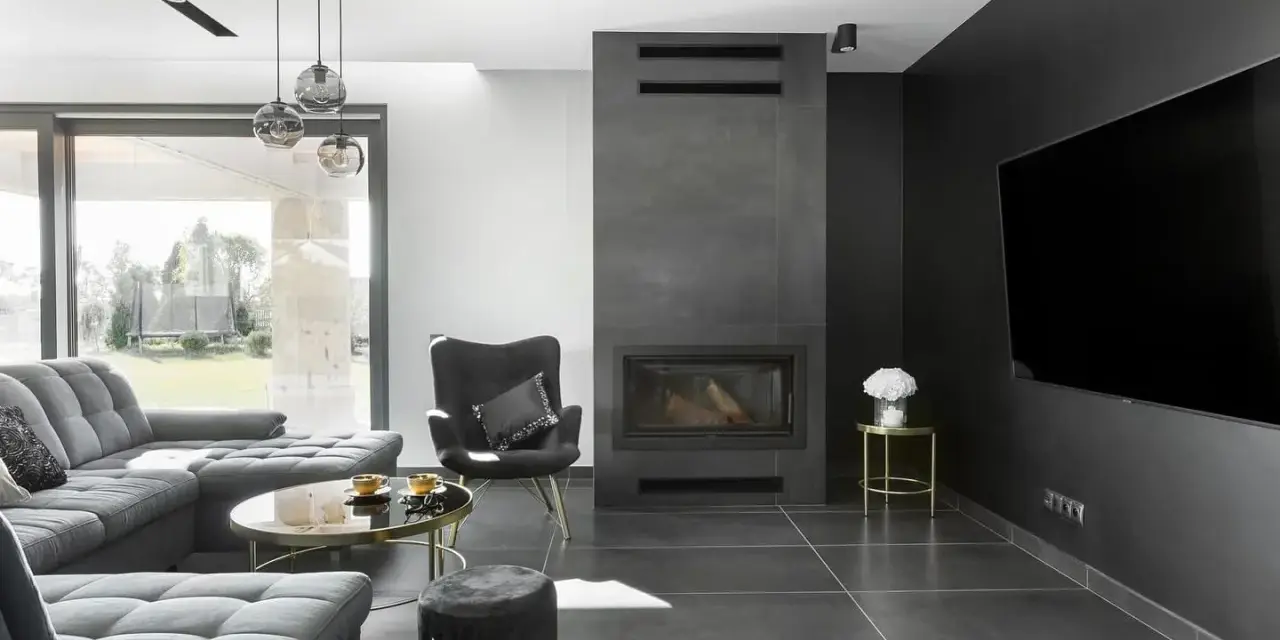 Nowoczesny salon z kominkiem inspiracje: szara sofa, czarny fotel, okrągły stolik kawowy i duży telewizor na ciemnej ścianie.