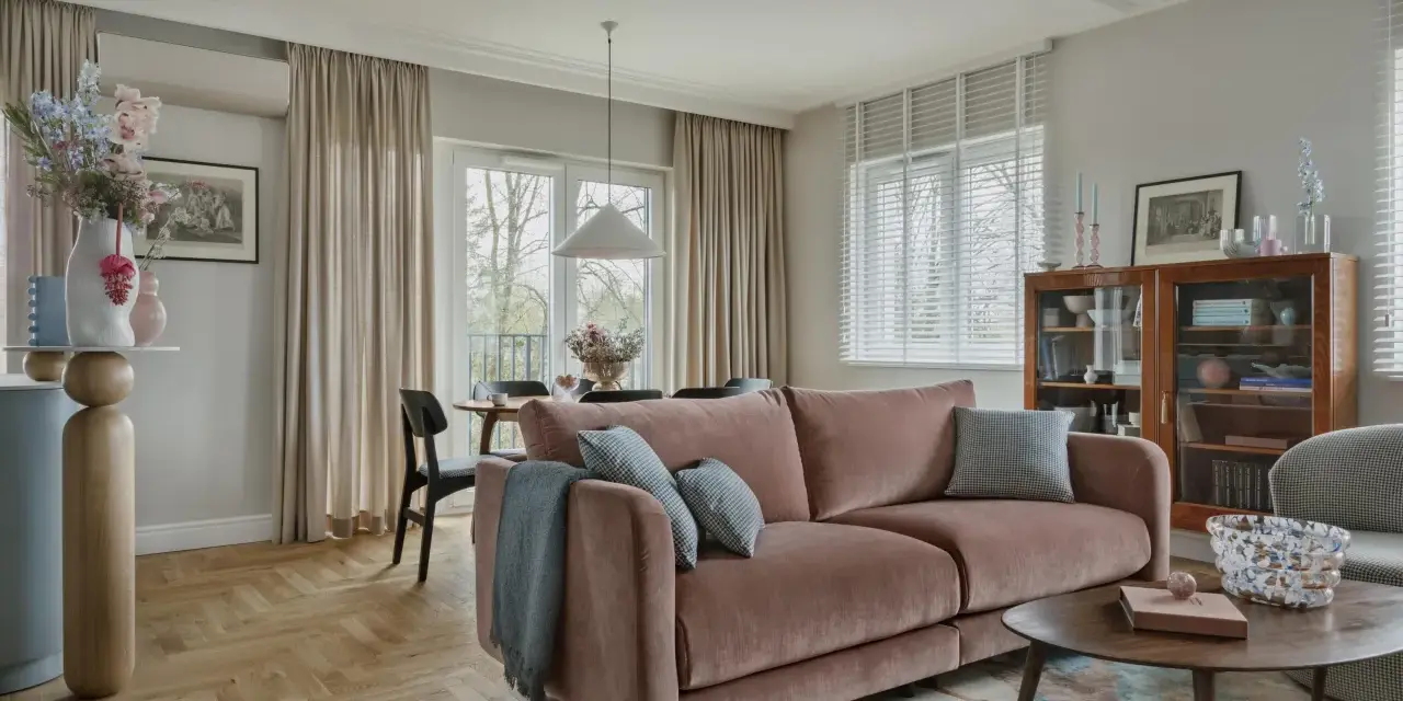 Przytulny salon z różową kanapą na środku, stolikiem kawowym, regałem i jadalnią w tle.