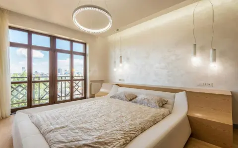 Beżowa sypialnia inspiracje: nowoczesne łóżko, okno z widokiem na miasto, okrągła lampa i subtelne oświetlenie tworzą przytulną atmosferę.