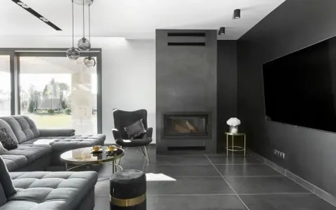 Nowoczesny salon z kominkiem inspiracje: szara sofa, czarny fotel, okrągły stolik kawowy i duży telewizor na ciemnej ścianie.
