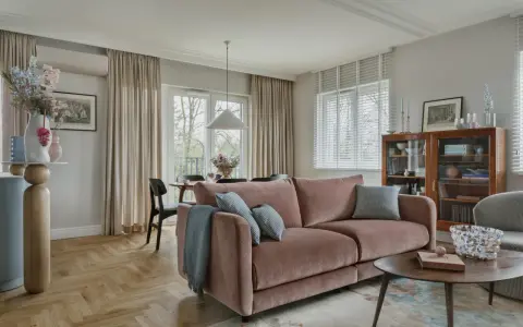 Przytulny salon z różową kanapą na środku, stolikiem kawowym, regałem i jadalnią w tle.