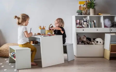 Dwie dziewczynki bawią się przy stoliku, tworząc przytulny kącik zabaw dla dziecka w salonie. Na półkach stoją pluszaki i książki.