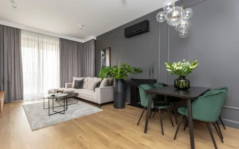 Nowoczesny salon z sofą w odcieniu beżu, dywanem w barwie szarej i zielonymi krzesłami przy czarnym stole.