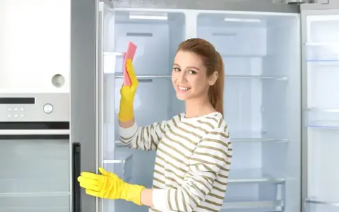 Kobieta w żółtych rękawiczkach czyści wnętrze lodówki różową gąbką. Pokazuje, jak myć lodówkę, by była lśniąca.