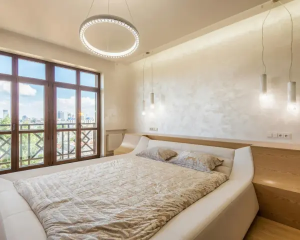 Beżowa sypialnia inspiracje: nowoczesne łóżko, okno z widokiem na miasto, okrągła lampa i subtelne oświetlenie tworzą przytulną atmosferę.