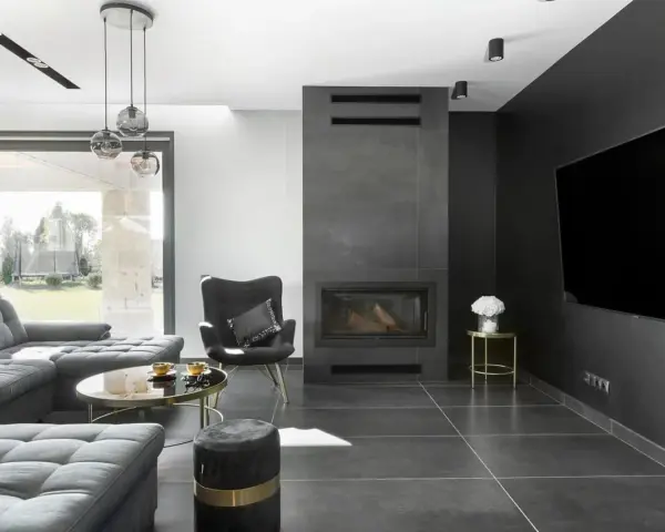 Nowoczesny salon z kominkiem inspiracje: szara sofa, czarny fotel, okrągły stolik kawowy i duży telewizor na ciemnej ścianie.