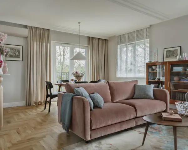 Przytulny salon z różową kanapą na środku, stolikiem kawowym, regałem i jadalnią w tle.