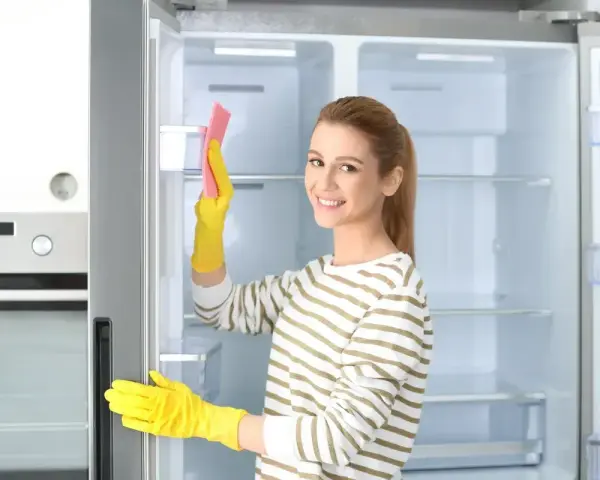 Kobieta w żółtych rękawiczkach czyści wnętrze lodówki różową gąbką. Pokazuje, jak myć lodówkę, by była lśniąca.