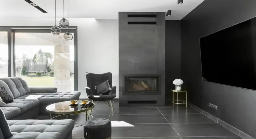 Nowoczesny salon z kominkiem inspiracje: szara sofa, czarny fotel, okrągły stolik kawowy i duży telewizor na ciemnej ścianie.
