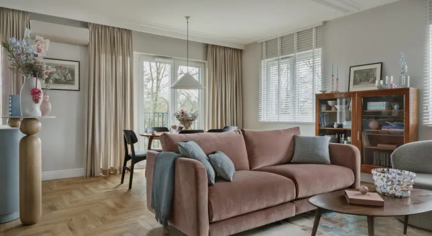 Przytulny salon z różową kanapą na środku, stolikiem kawowym, regałem i jadalnią w tle.