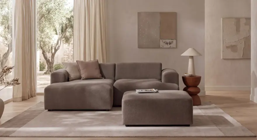 Wygodna sofa i puf w kolorze greige jaki to kolor, idealnie komponują się z jasnym wnętrzem i widokiem na drzewa.