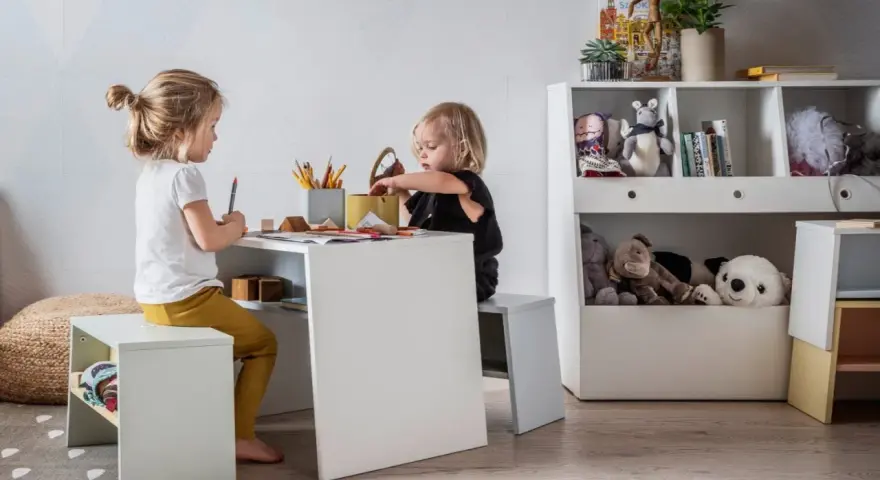 Dwie dziewczynki bawią się przy stoliku, tworząc przytulny kącik zabaw dla dziecka w salonie. Na półkach stoją pluszaki i książki.