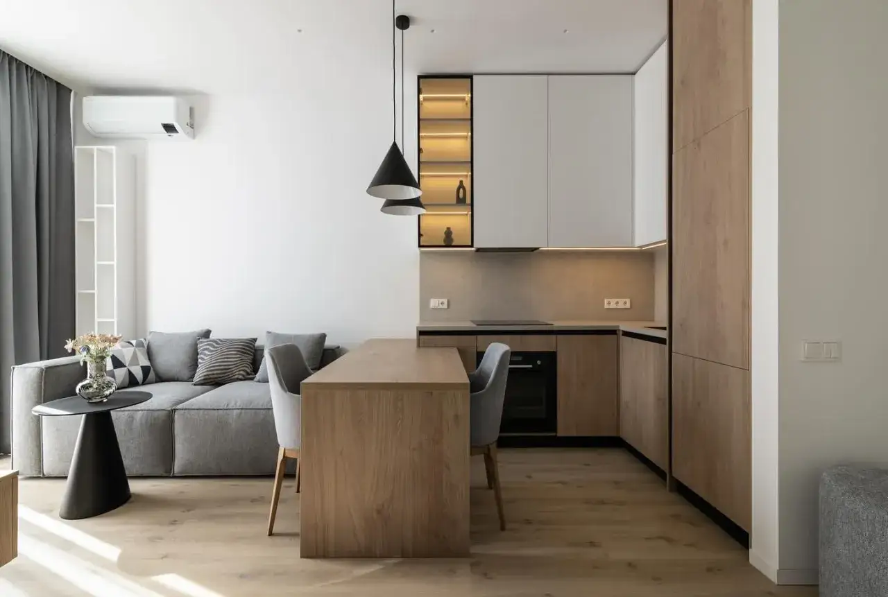 Nowoczesny salon 16m2 aranżacja: szara sofa, drewniany st&oacute;ł z krzesłami, aneks kuchenny z białymi i drewnianymi frontami.