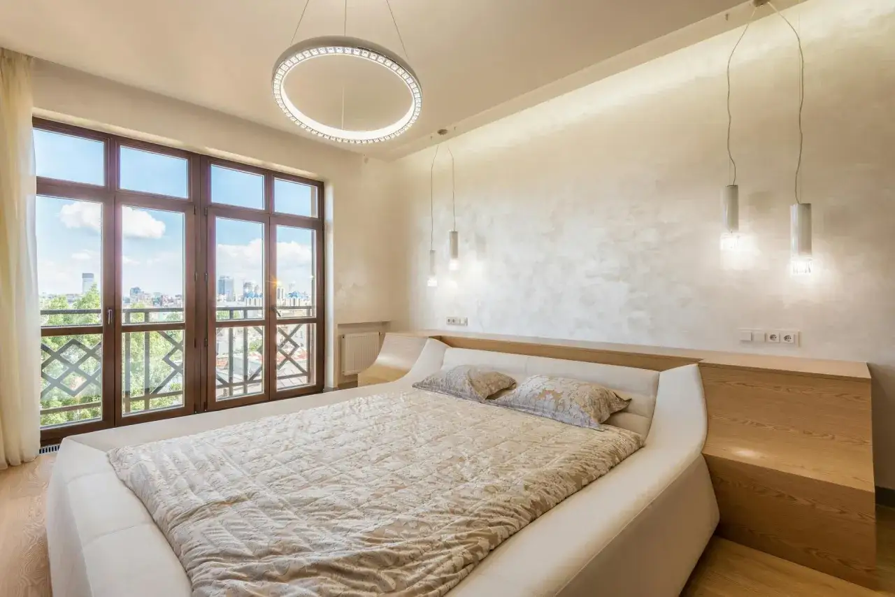 Beżowa sypialnia inspiracje: nowoczesne ł&oacute;żko, okna z widokiem na miasto, designerskie oświetlenie.