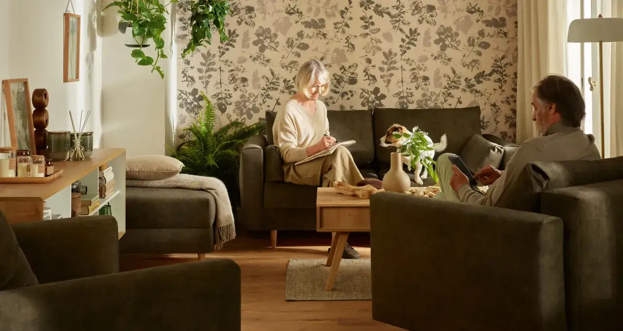 Przytulny salon z kobietą piszącą w notatniku i mężczyzną korzystającym z tabletu. Idealne inspiracje, jak urządzić pok&oacute;j dla babci.