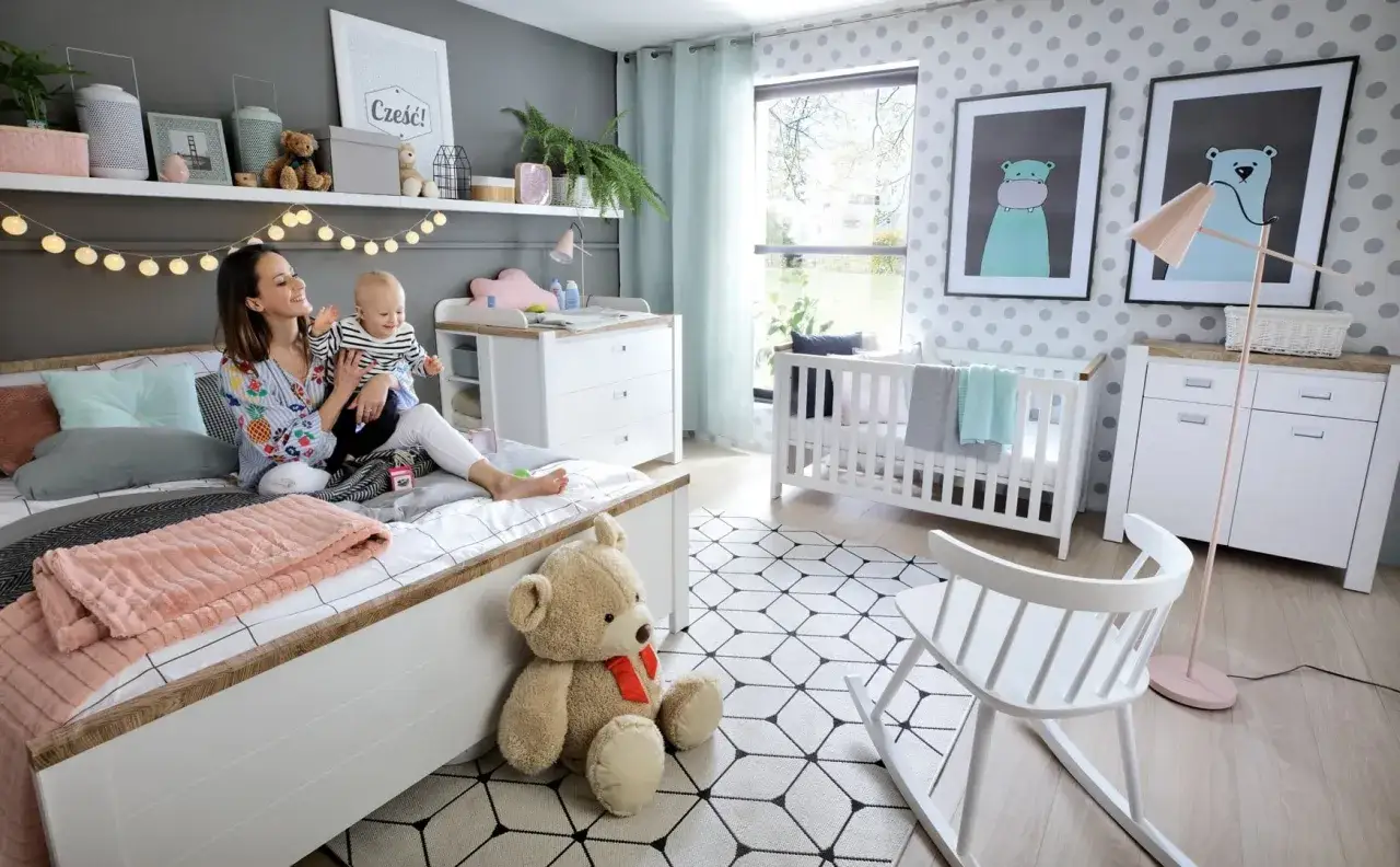 Mama i dziecko cieszą się wsp&oacute;lnym pokojem rodzic&oacute;w z dzieckiem. Na ł&oacute;żku siedzi uśmiechnięta kobieta z niemowlęciem na rękach.