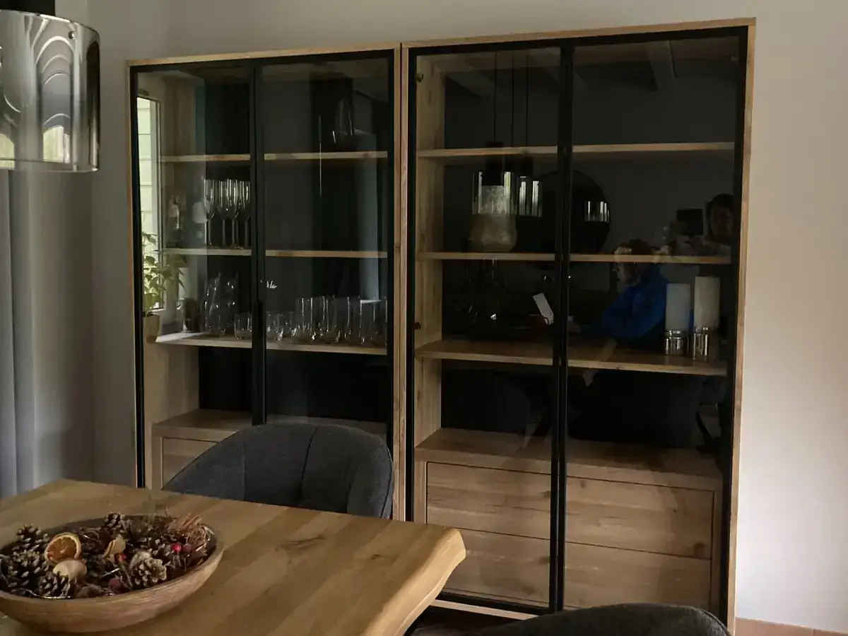 Drewniana witryna z przeszklonymi drzwiczkami, idealna do salonu. W środku kieliszki, dzbanki i dekoracje.