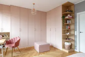 Jak urządzić pokój dzienny z szafą? Jasnoróżowa zabudowa, musztardowy fotel i drewniane półki tworzą przytulną przestrzeń.